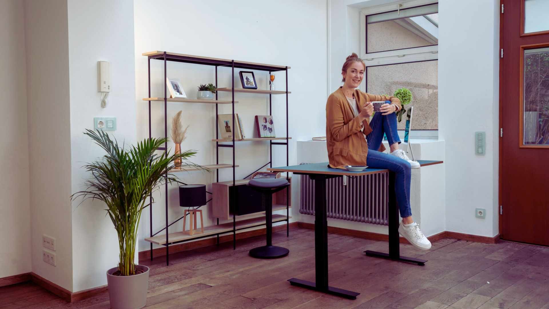 Eine Frau sitzt im Büro auf dem Schreibtisch mit Kaffee in der Hand.